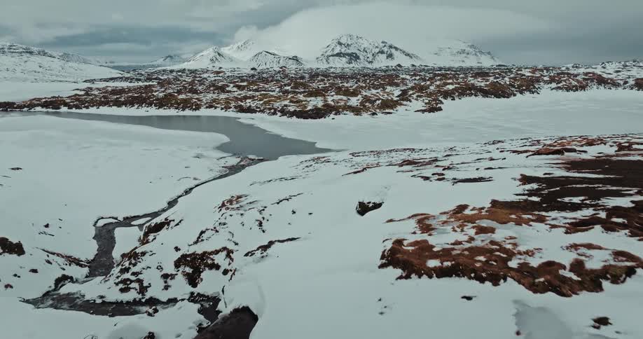 An aerial flight over a vast, frozen Icelandic landscape reveals a winding river and snow-covered mountains under a cloudy sky