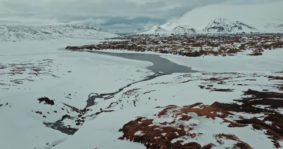 An aerial view slowly glides over a vast, desolate, snow-covered landscape revealing a frozen lake and majestic mountains in the distance