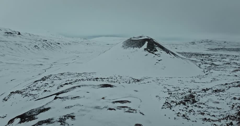 A slow aerial drone shot pushes forward towards a dormant volcano blanketed in snow in a vast and desolate winter landscape