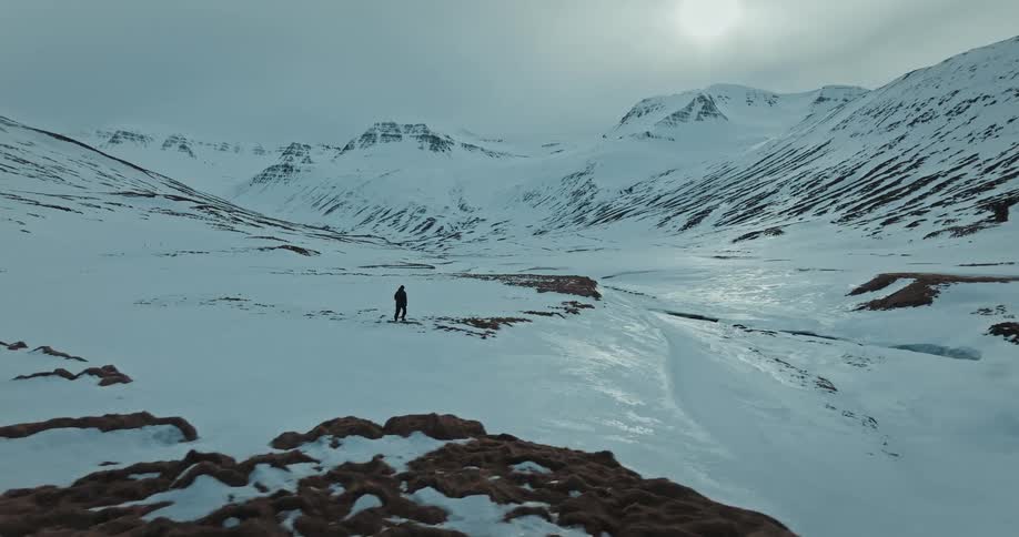 An aerial shot follows a lone hiker as they traverse a vast, snow-covered valley surrounded by majestic mountains under a winter sun