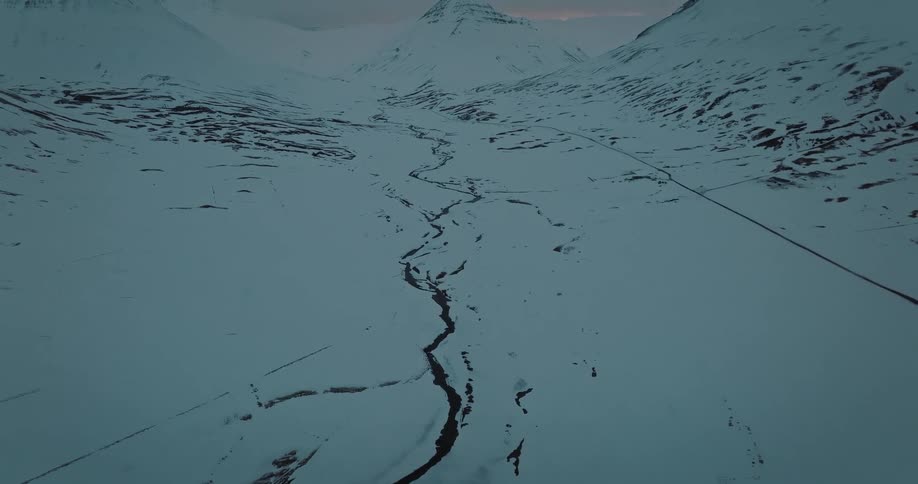 An epic aerial view pulls back to reveal a vast, desolate snow-covered valley with a winding river in the Icelandic highlands at dusk
