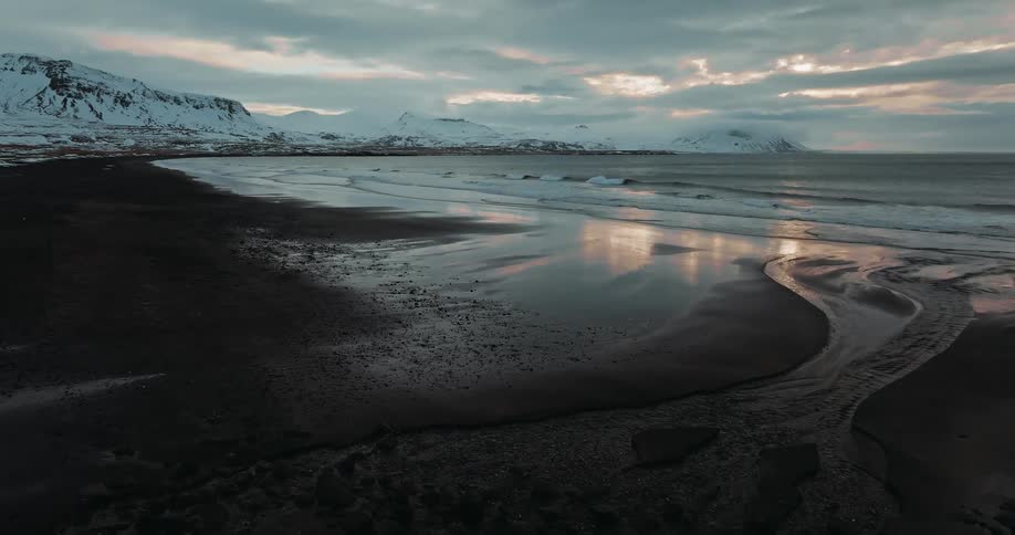 Stunning aerial view slowly pushes forward over a snowy hill to reveal the vast, reflective black sand beach of an Icelandic coastline at sunset