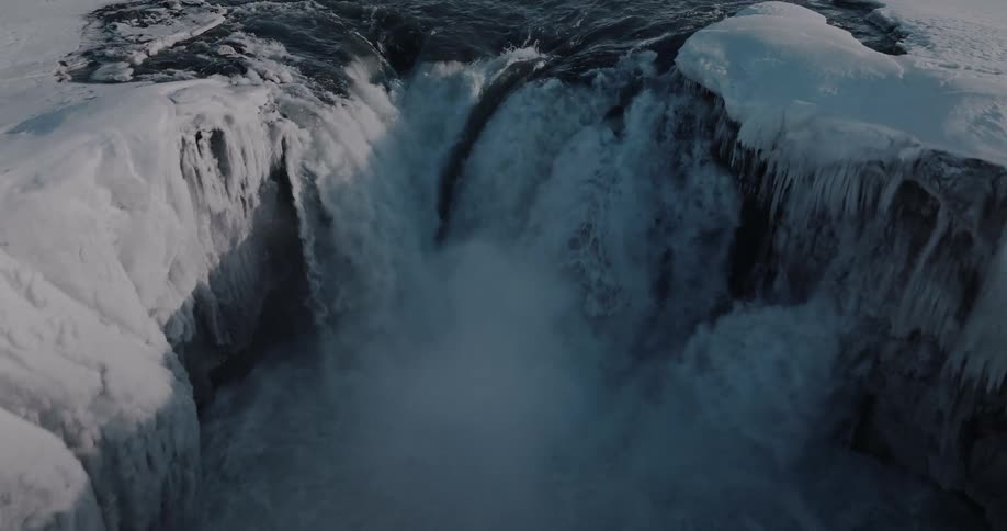 An aerial view pulls back from turbulent dark water to reveal a powerful, icy waterfall crashing through a frozen winter canyon