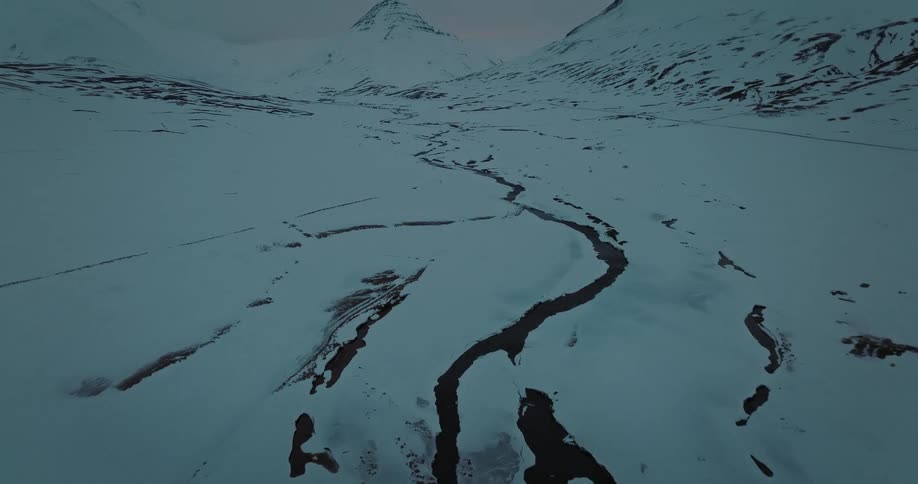 A cinematic aerial view slowly tilts up from a dark, winding river to reveal a vast, snow-covered mountain valley at twilight