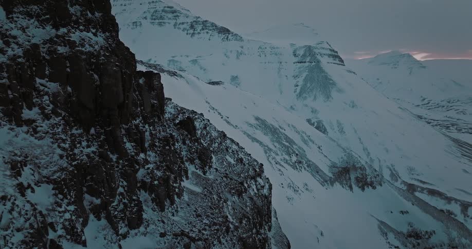 An aerial shot slowly pulls back along a rocky cliff to reveal a vast, snow-covered mountain valley at dusk