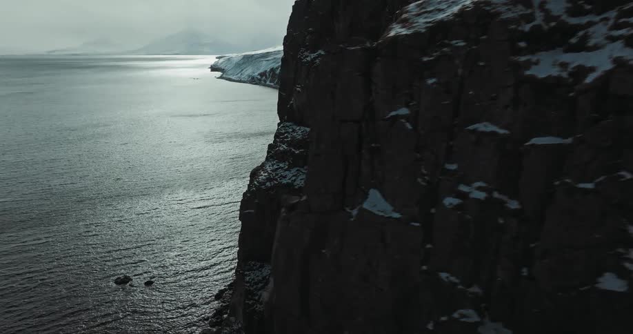 A cinematic aerial flight reveals a majestic and rugged winter coastline with snow-covered cliffs and a dark ocean