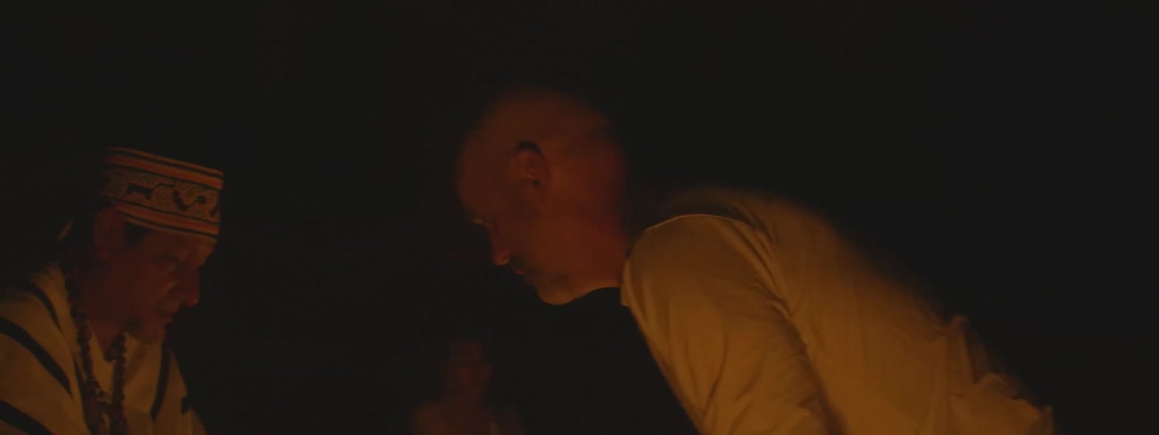 A shaman guides a man through a sacred candlelight ritual in a dark, intimate setting