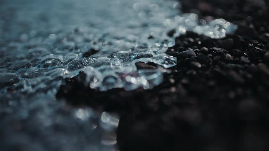 Close up slow motion shot of a thin sheet of ice melting over dark gravel