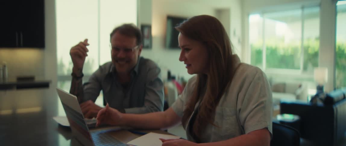 A woman explains something on a laptop to a smiling man as they collaborate on a project from their home kitchen