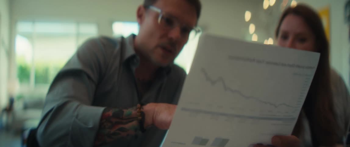 A businessman with glasses and tattoos explains a declining financial chart to a client during a meeting