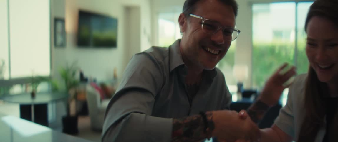 A tattooed man and a woman joyfully shake hands and celebrate a successful agreement in a sunny, modern living room