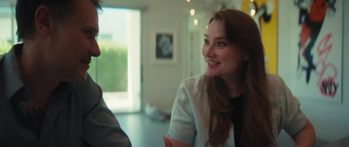 A man and woman playfully arm wrestle across a table in a modern home, both laughing and smiling