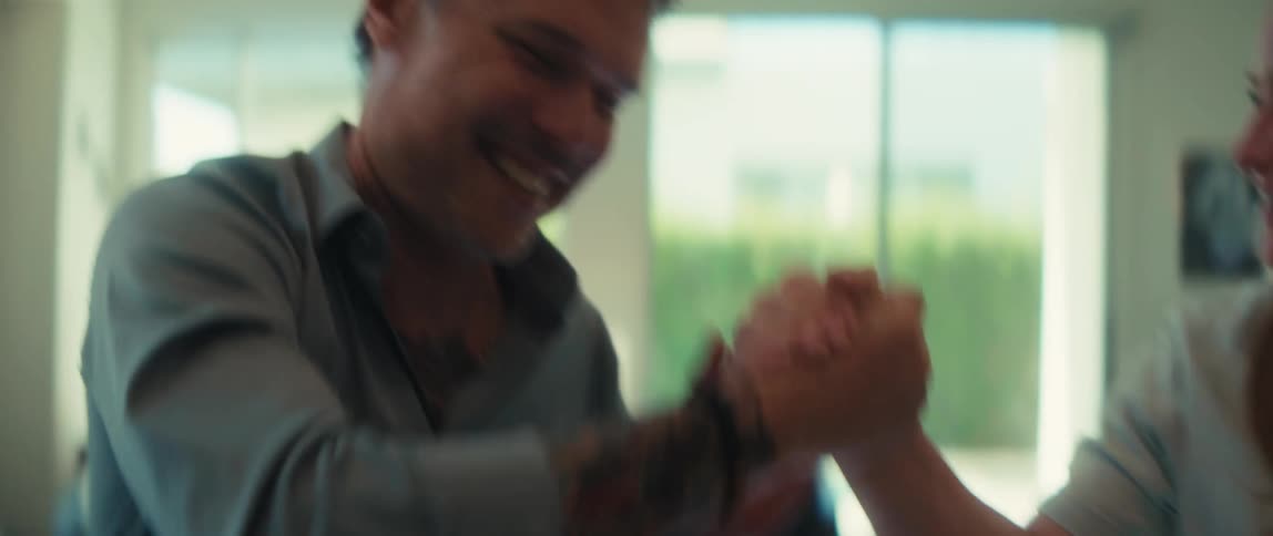 A smiling man with tattoos playfully arm wrestles another person in a sunlit room
