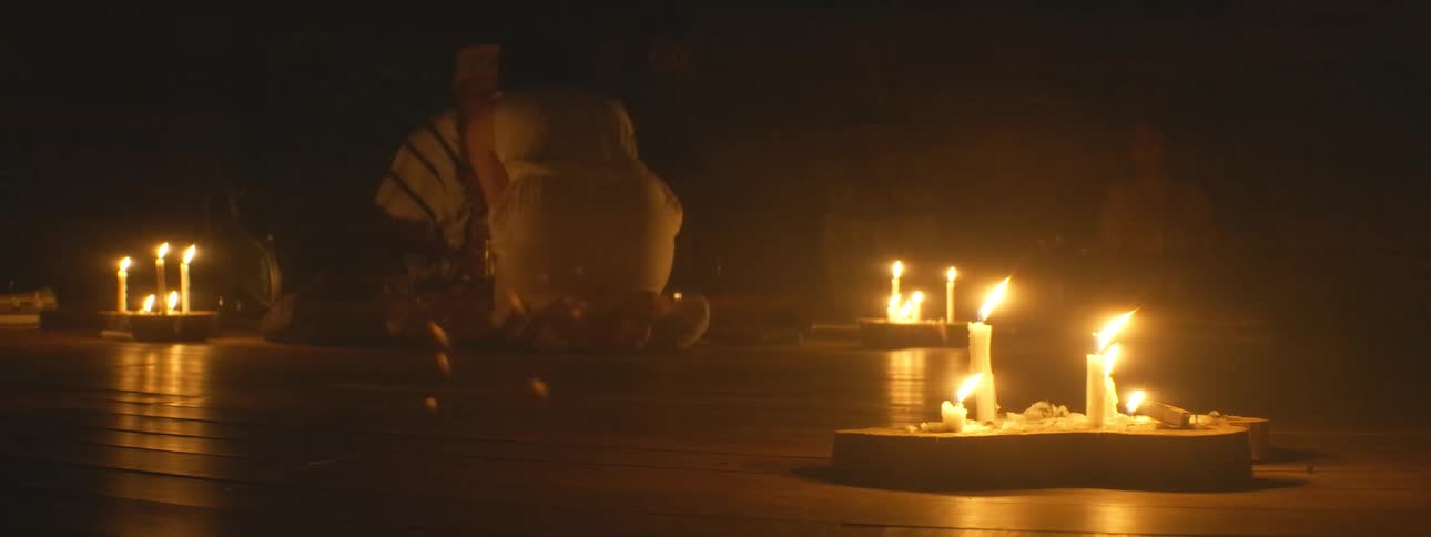 A traditional indigenous ceremony unfolds in a dark, hazy room illuminated only by flickering candles
