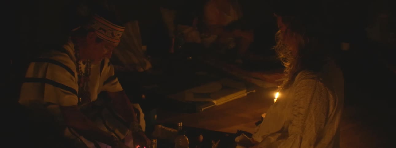 A shaman performs a spiritual healing ceremony for a woman in a dark, candlelit room with a group of people