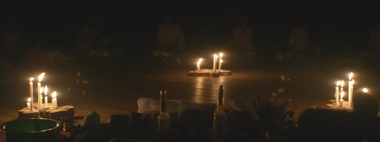A mysterious nighttime spiritual ceremony takes place in a dark room with people meditating in a circle around flickering candles