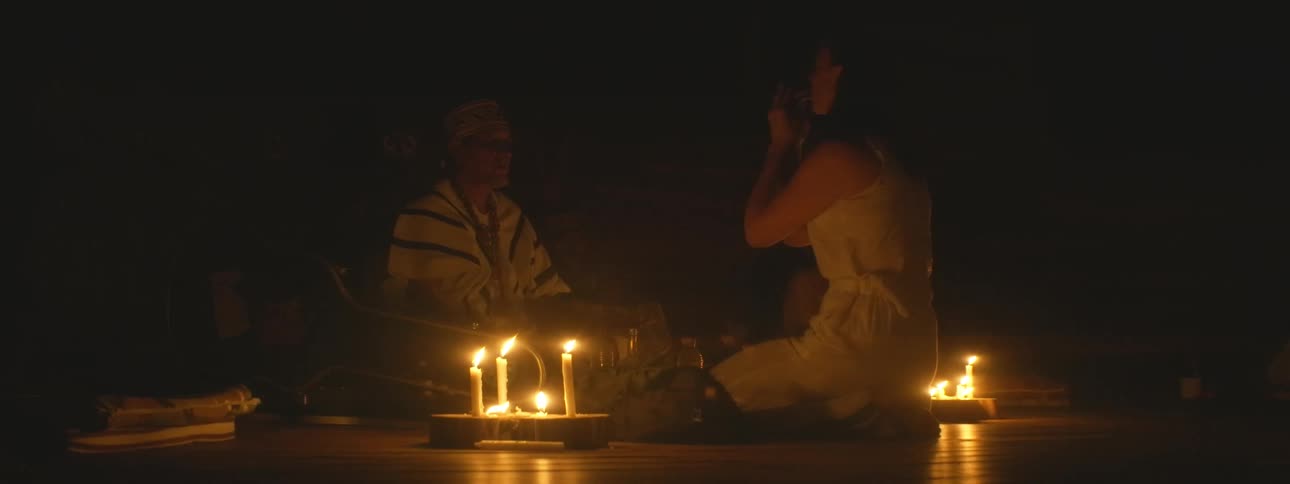 A shaman leads a woman through a sacred healing ceremony in a dark room illuminated only by flickering candles