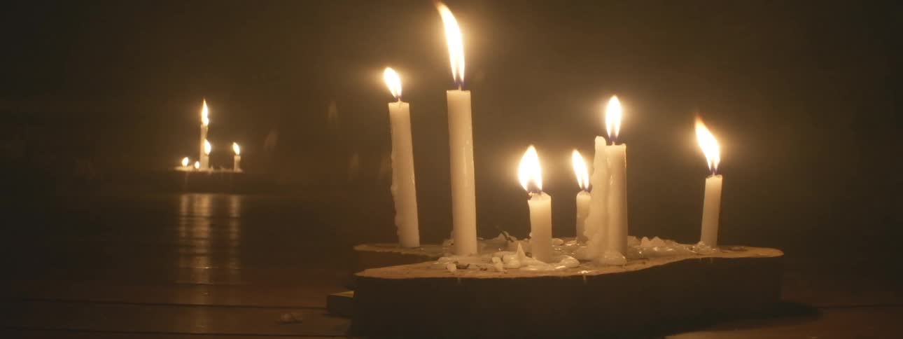 Multiple white candles with flickering flames are arranged on the floor, illuminating a dark, hazy room