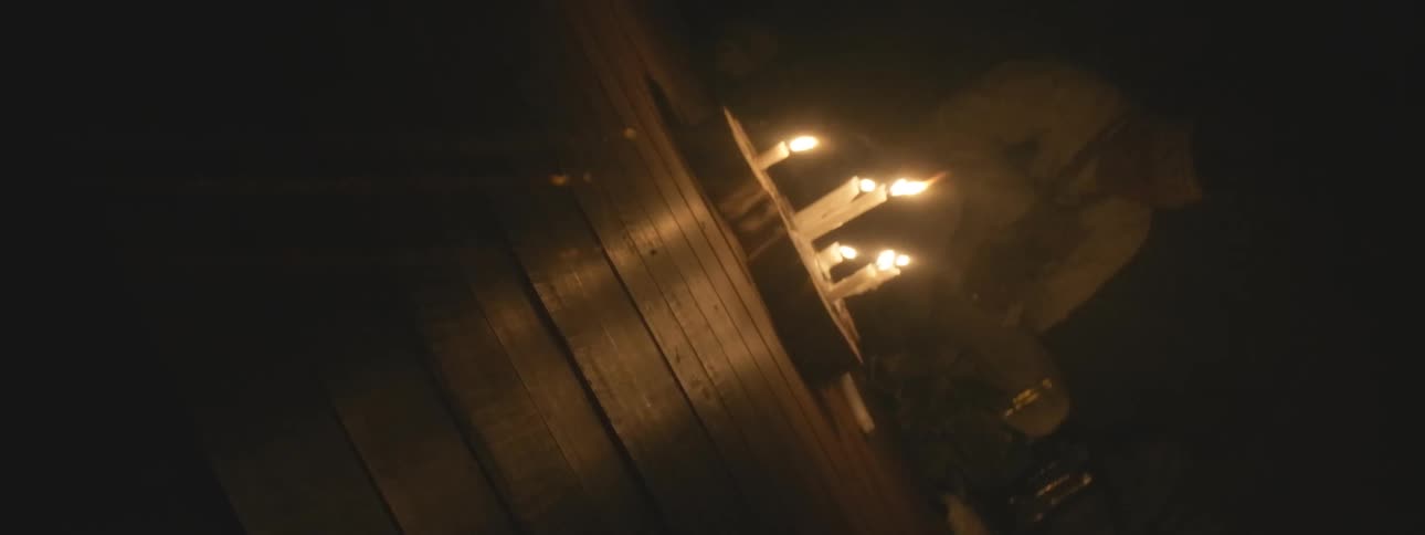A mysterious figure in traditional garb kneels by flickering candlelight in a dark wooden room