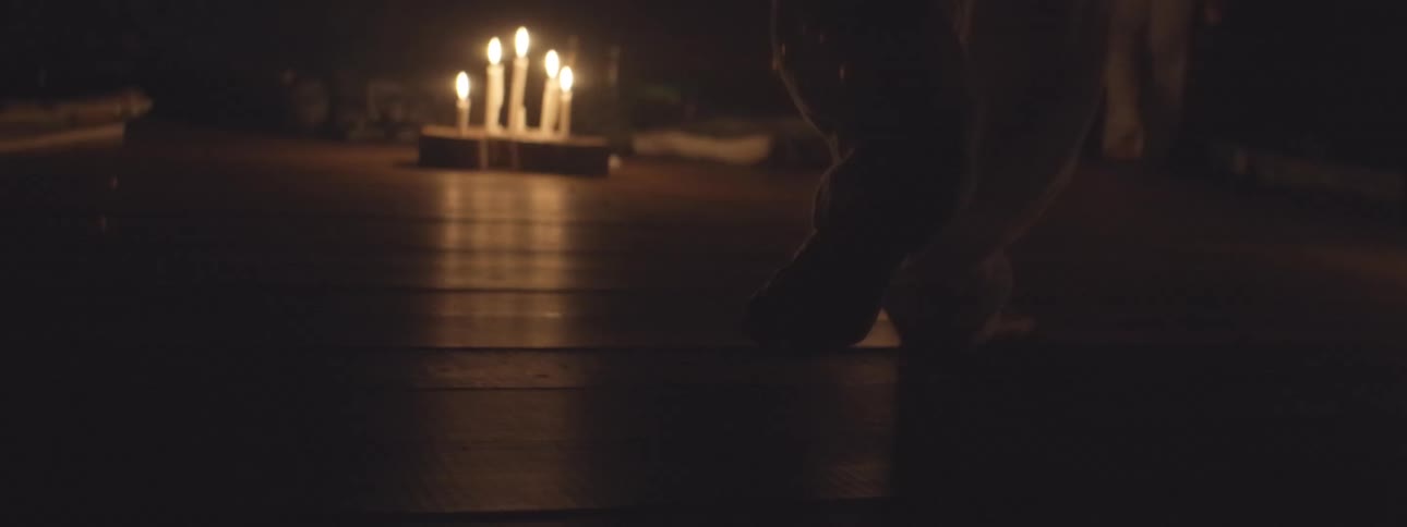 Slow-motion close-up of a person's bare feet walking across a wooden floor in a dark, candlelit room