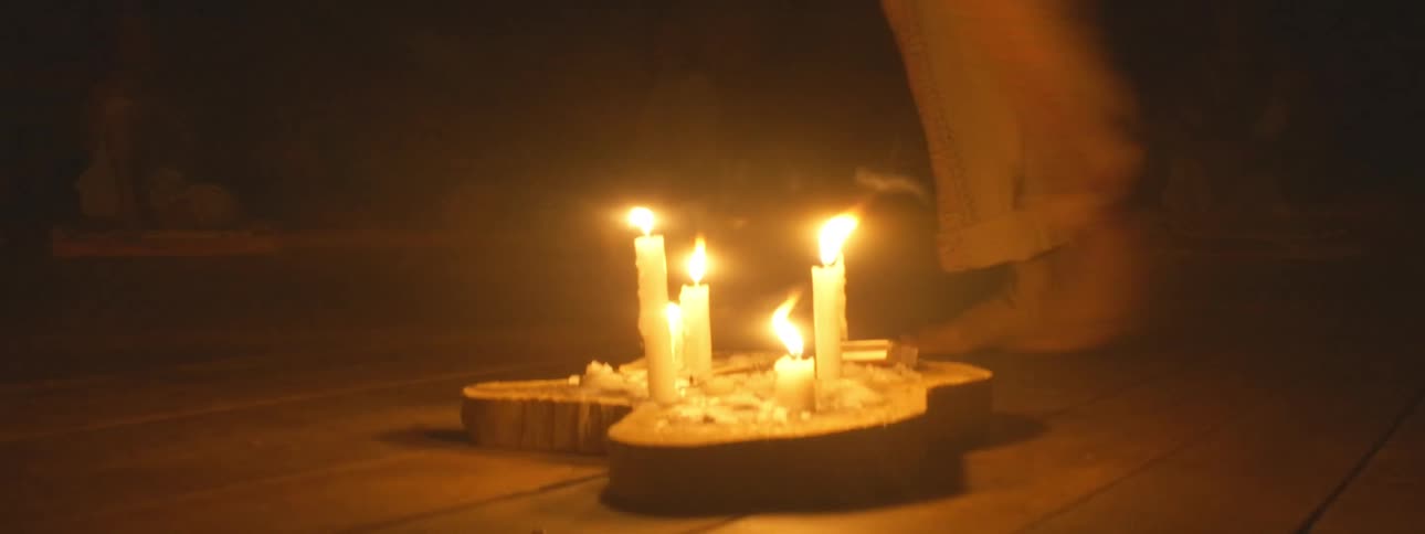 A person performs a barefoot ritual, walking around lit candles in a dark, spiritual ceremony