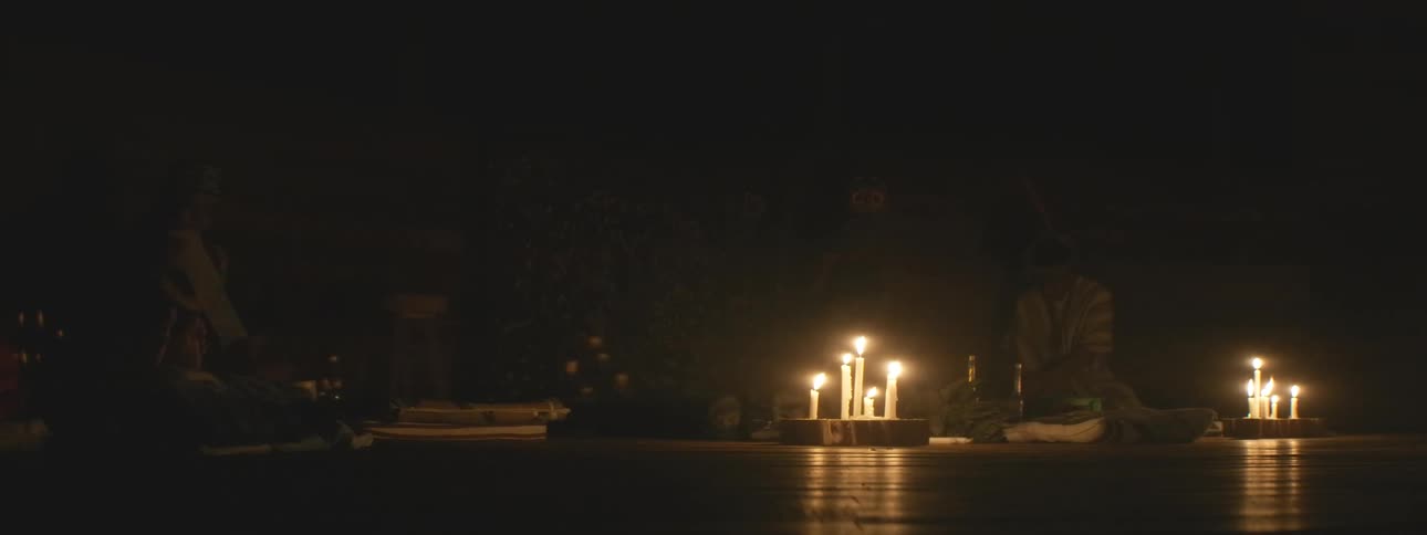 A shaman sits on the wooden floor of a dark room, performing a spiritual healing ceremony illuminated by flickering candles