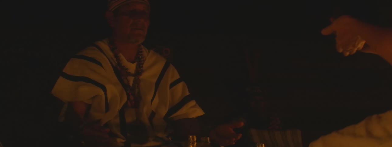 An indigenous shaman in traditional attire performs a sacred candlelit ritual, placing a small item into another person's cupped hands