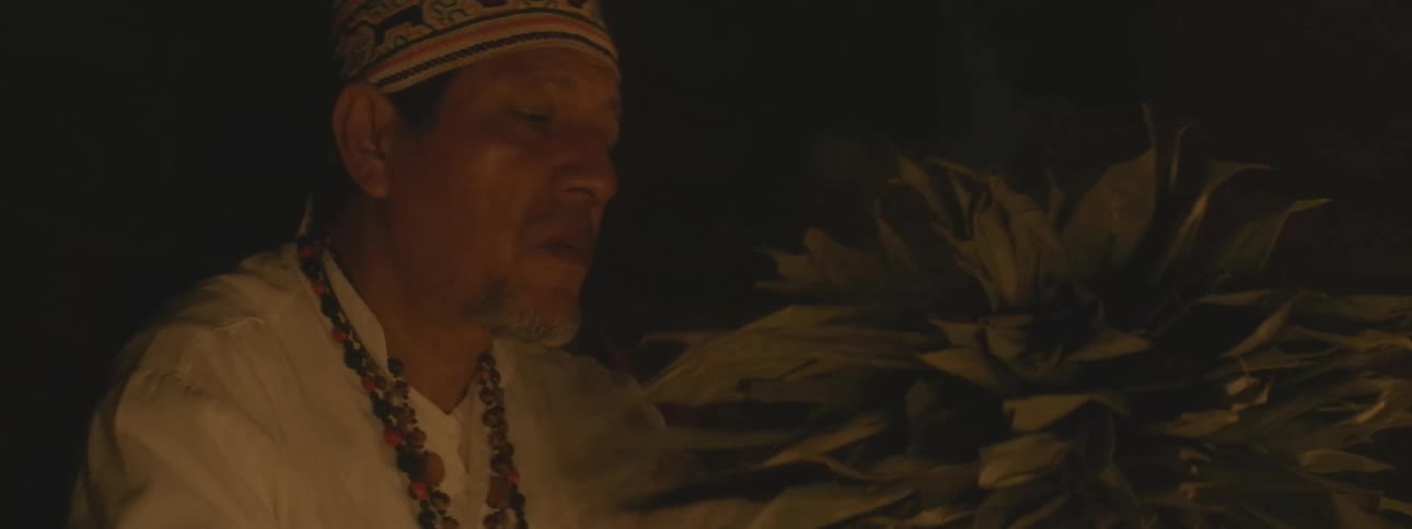 A shaman in traditional clothing performs a spiritual cleansing ritual by blowing cigarette smoke over a bundle of leaves by candlelight