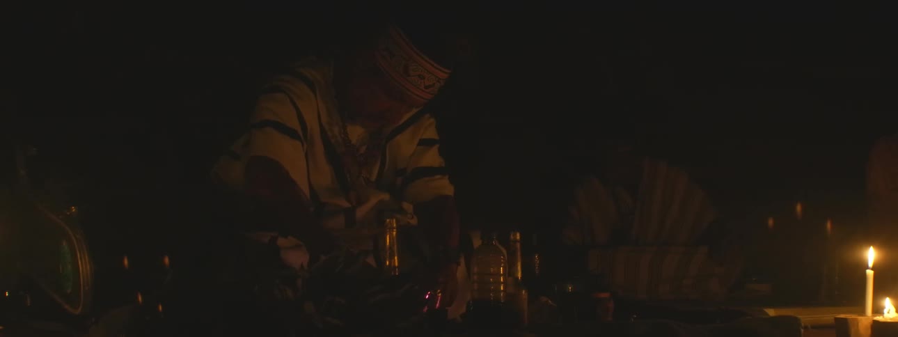 A shaman in traditional attire performs a sacred candlelit ceremony in a dark room, preparing ritualistic elements