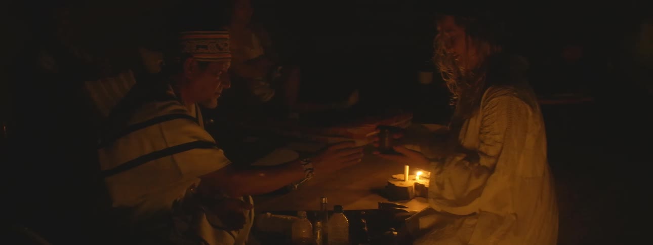 A woman partakes in a traditional shamanic ritual, drinking a ceremonial brew offered by a shaman in a room lit by candlelight