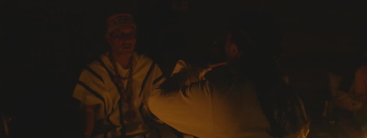 An indigenous shaman in traditional attire performs a sacred candlelit ritual, offering a ceremonial drink and a blessing to a participant