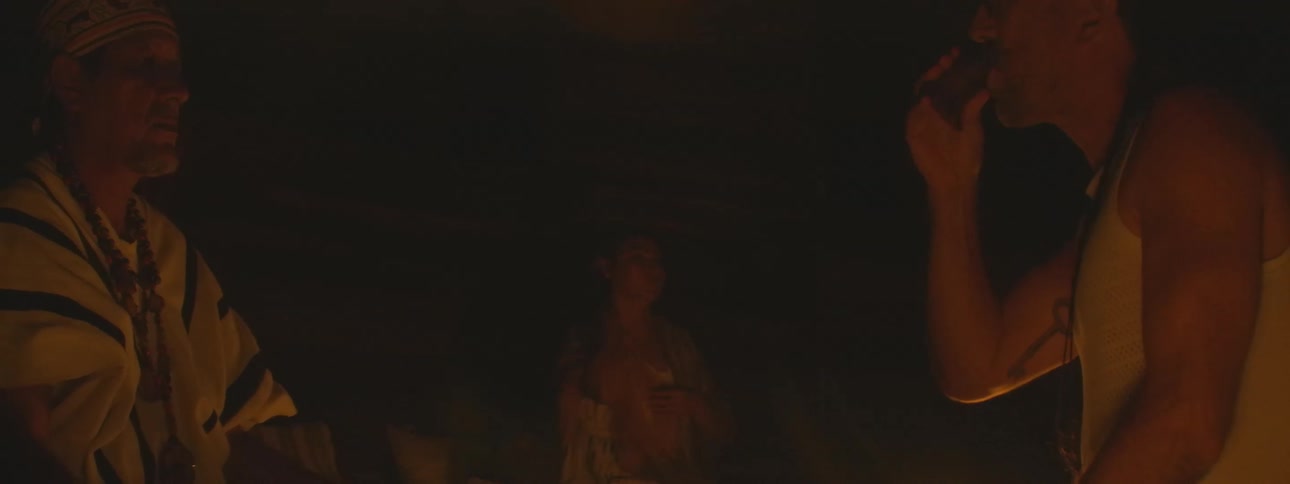 A shaman leads a traditional healing ceremony by candlelight, serving a sacred brew to a participant in a dark room