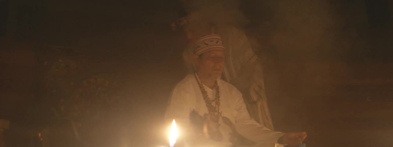 An indigenous shaman leads a spiritual purification ritual with smoking incense and candles in a dark, sacred setting