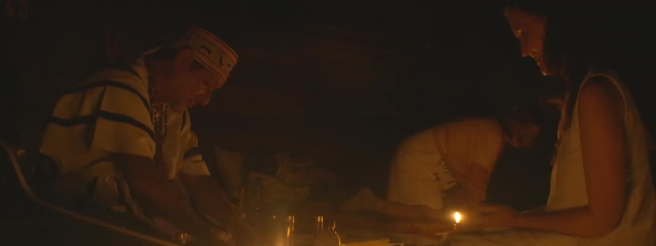 A shaman and two women participate in a traditional spiritual healing ceremony in a dark room illuminated by candlelight