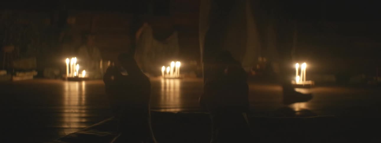 A low-angle view from a prone position captures a mystical candlelit dance performance by silhouetted figures in a dark room