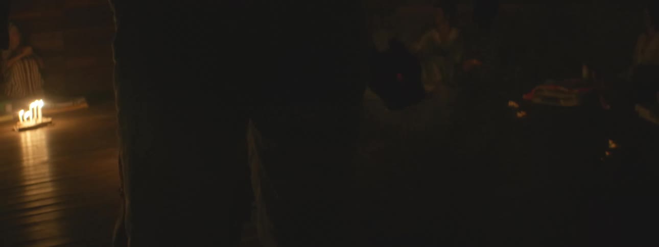 A cloaked figure stands in a dark, candlelit room as smoke drifts through the air during a mysterious ceremony