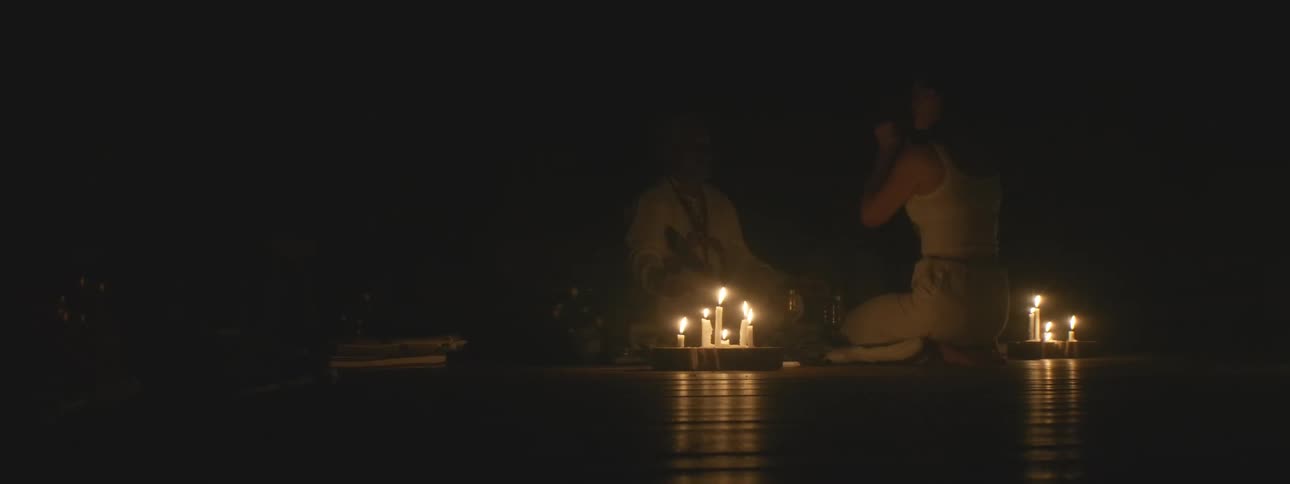 A shaman and a woman participate in a sacred spiritual ceremony illuminated by candlelight in a dark, tranquil space