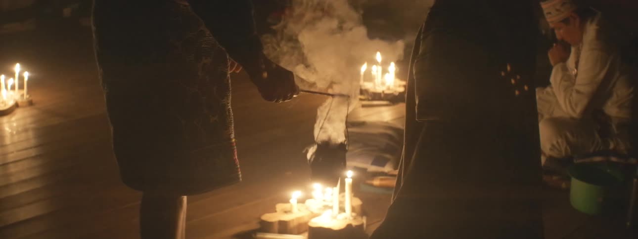 A shaman performs a spiritual cleansing ritual with smoke from a censer in a dark room lit by candles