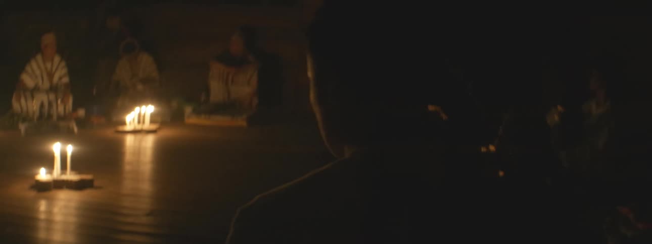 A shaman leads a spiritual healing ceremony in a dark room illuminated by the warm glow of candles