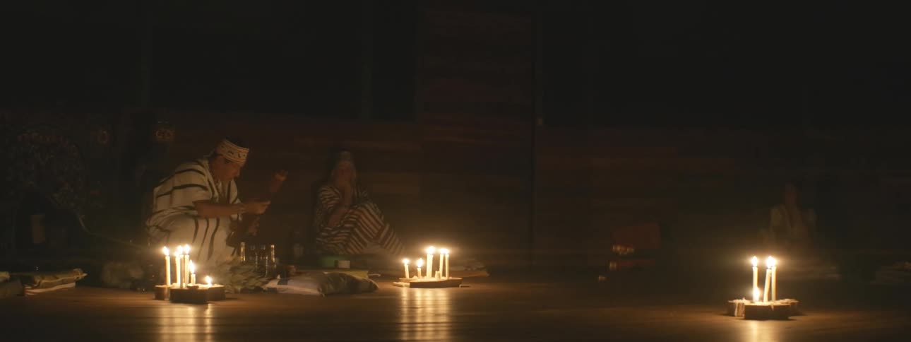 A shaman prepares for a spiritual ritual in a dark, candlelit room, accompanied by an assistant