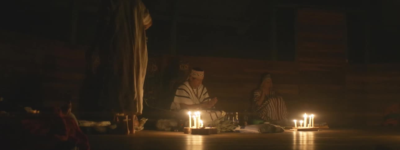 A group of indigenous people in traditional attire perform a sacred spiritual ceremony by candlelight in a dark room