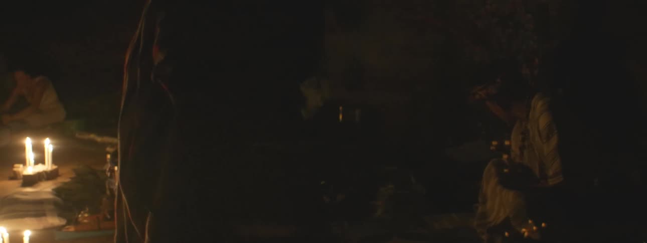 A mystical shamanic ceremony takes place at night, illuminated by candlelight, as a spiritual leader waves a smoking censer