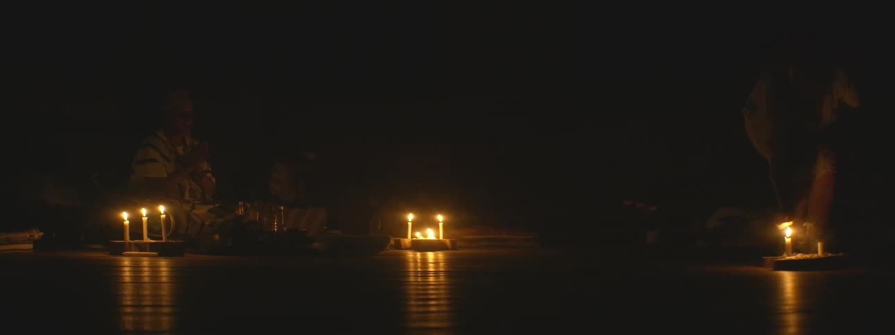 A group of people participate in a mysterious shamanic ritual, chanting and dancing in a dark room illuminated only by candlelight