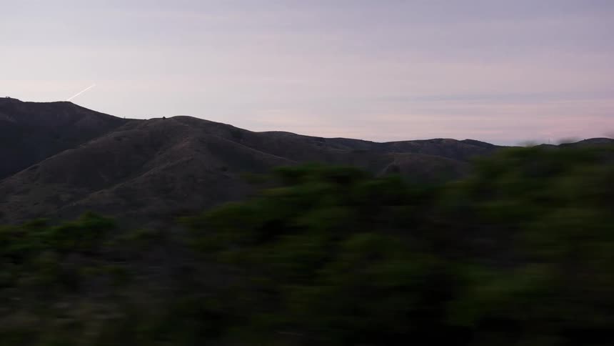A scenic side-view tracking shot from a vehicle driving past rolling hills and mountains at dusk