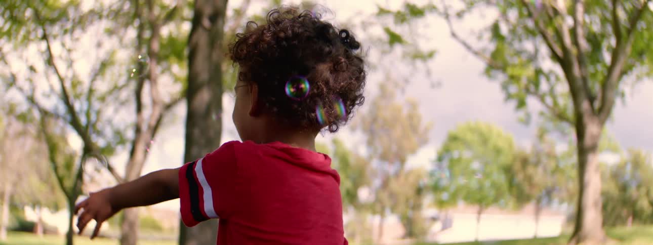 A joyful toddler in a red shirt laughs as bubbles float around him in a sunny park in slow motion