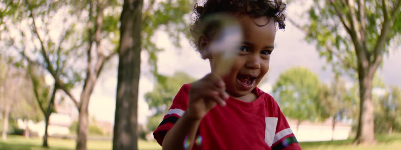 Slow motion shot of an adorable little boy in a red shirt laughing as he plays with soap bubbles in a park