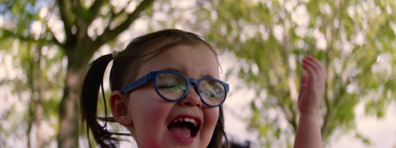 A delighted little girl with glasses laughs and plays with bubbles outdoors with a friend in slow motion