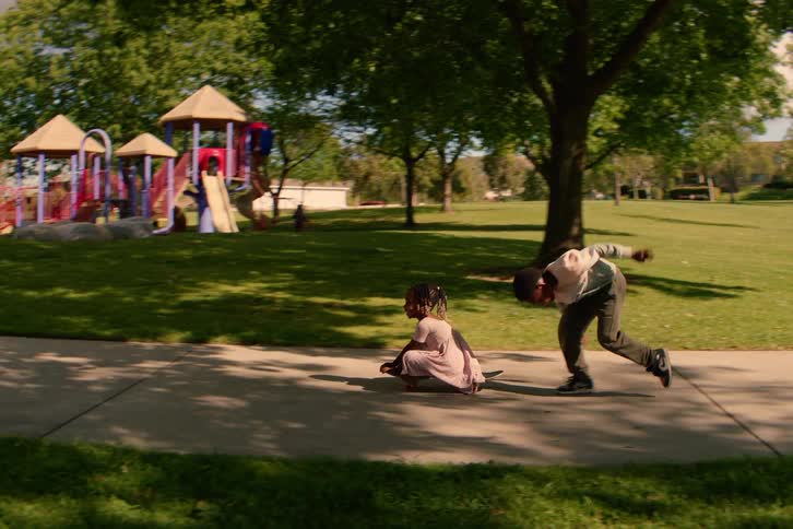 An older brother pushes his younger sister on a skateboard down a path in a park in slow motion