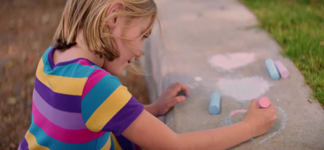 A creative young girl in a colorful striped shirt enjoys drawing hearts with sidewalk chalk on a concrete step