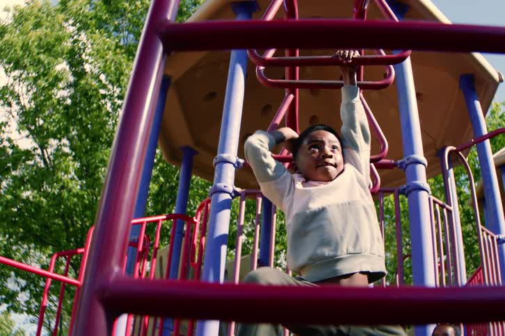 A young boy proudly hangs from the monkey bars on a colorful playground while his little sister watches from below